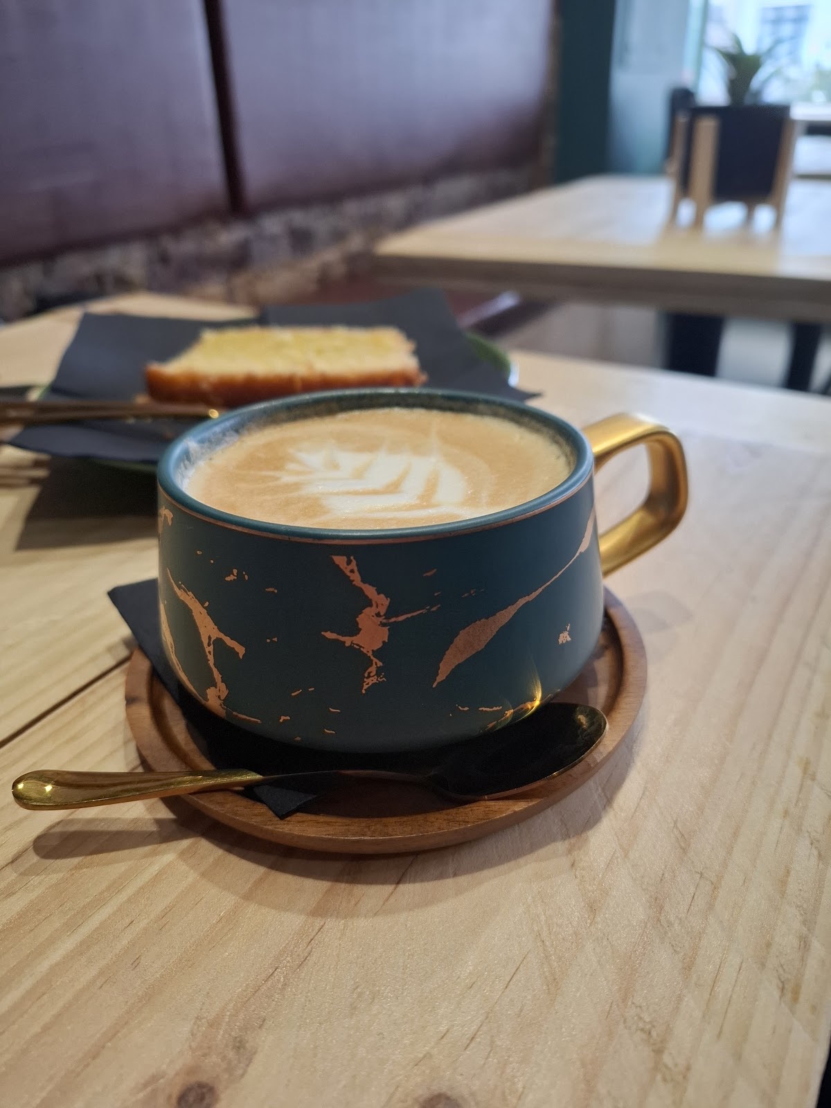 A wide teal and gold cup of latte with leaf art beside a slice of toasted cake